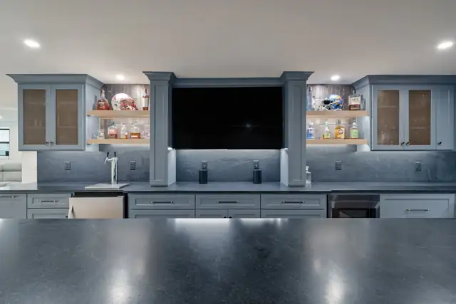 Finished basement bar in the Kansas City area with blue-gray cabinetry, honed black counters, under-cabinet lighting and a wall-mounted TV