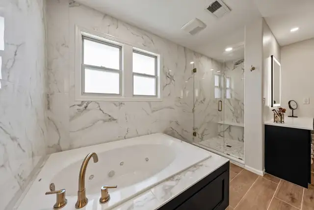Primary bathroom with a jetted tub, marble-look tile walls, frameless glass shower and black vanity