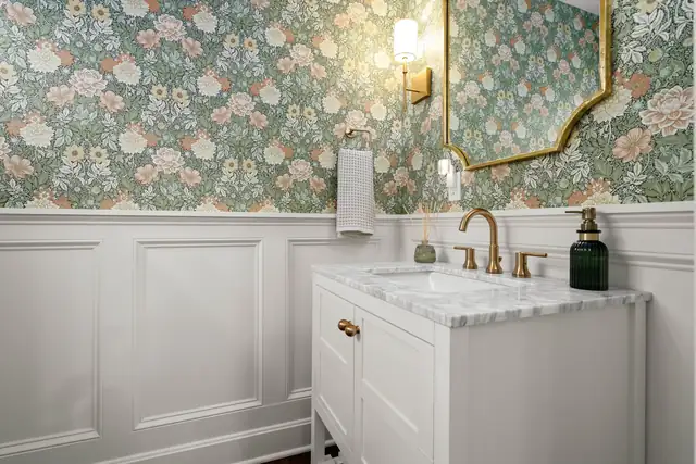 Powder room with floral wallpaper, white wainscoting, marble-top vanity and brass faucet